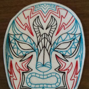 Puede incluir: Una máscara de fieltro blanco con un diseño bordado en negro y azul. La máscara presenta una cara estilizada con una nariz grande, ojos y boca.