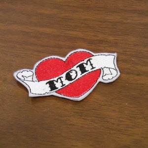 May include: White felt patch with a red heart and the word "MOM" in black lettering. The heart is surrounded by a white banner.