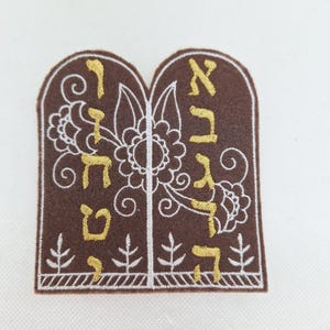 Op de afbeelding: Een bruine vilt patch geborduurd met gouden Hebreeuwse letters en witte bloemen- en bladeren ontwerpen. De patch is gevormd als twee tabletten en heeft ingewikkelde stiksels en decoratieve elementen.