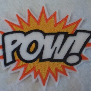 May include: A white embroidered patch with the word "POW!" in black outlined letters. The word is surrounded by a yellow and orange starburst.