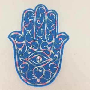 Puede incluir: Un parche azul con forma de mano de Hamsa con intrincados detalles bordados. El diseño presenta un símbolo de ojo central y patrones arremolinados en blanco, rosa y azul. El parche es un artículo decorativo.