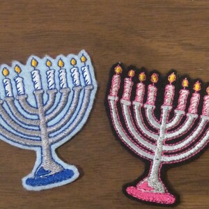 May include: Two embroidered iron menorah patches. One is blue with silver candles and the other is black with pink candles.