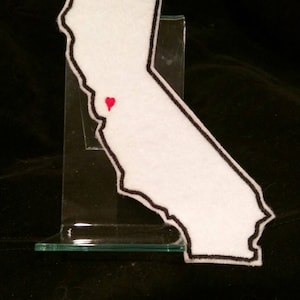 May include: White felt outline of the state of California with a red heart in the center, representing the location of San Francisco. The outline is sewn onto a white fabric background.