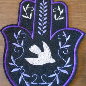 May include: A black felt Hamsa hand with a white dove in the center. The Hamsa is outlined in purple and has purple embroidered vines around the edges.