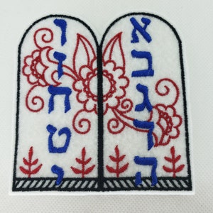 Op de afbeelding: Twee witte stoffen tabletten met blauwe en rode borduursels. De tabletten zijn aan elkaar verbonden en bevatten Hebreeuwse letters en bloemmotieven.