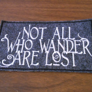Puede incluir: Un parche de fieltro gris con texto bordado en blanco que dice "Not all who wander are lost."