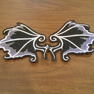 May include: A pair of black and purple embroidered wings with white accents. The wings are made of fabric and have a glittery finish.