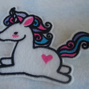 Puede incluir: Aplique de fieltro blanco de un unicornio de dibujos animados con un corazón rosa en un lado. El unicornio tiene una melena y una cola de color morado, azul y rosa.