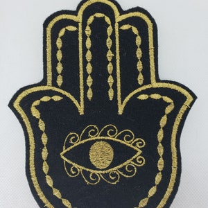 May include: A black felt hamsa hand with a gold embroidered outline and a gold eye in the center. The eye is surrounded by gold swirls.