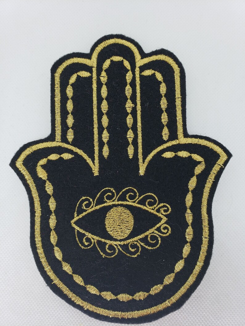 May include: A black felt hamsa hand with a gold embroidered outline and a gold eye in the center. The eye is surrounded by gold swirls.