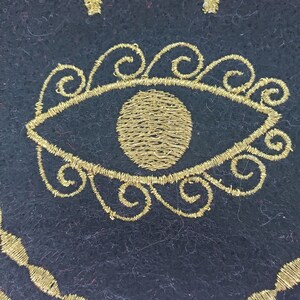 May include: A black felt circle with a gold embroidered eye design. The eye is surrounded by a circle of gold swirls.
