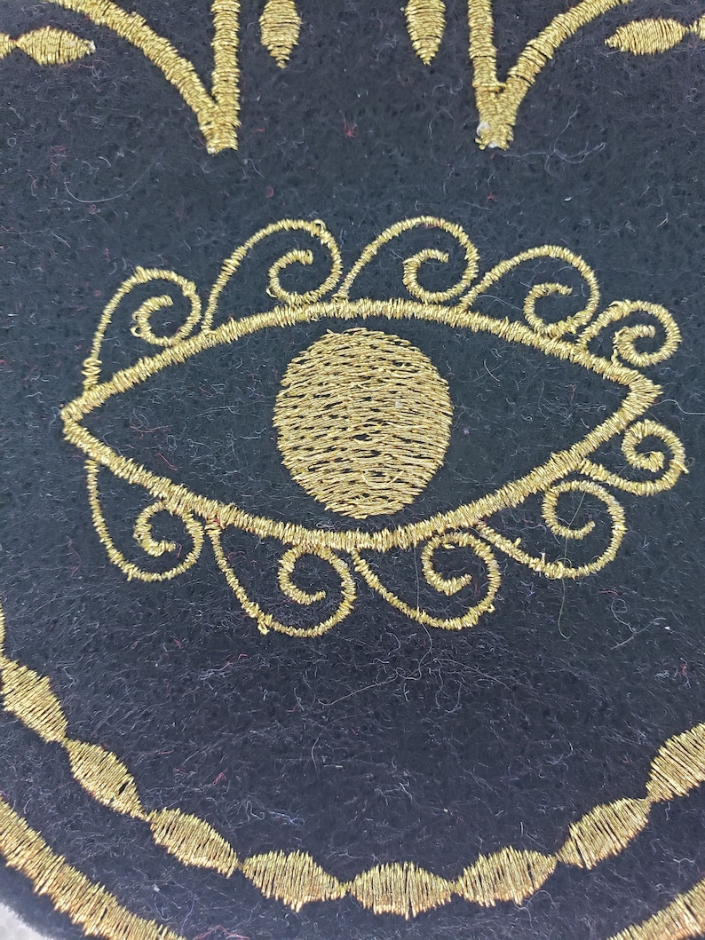 May include: A black felt circle with a gold embroidered eye design. The eye is surrounded by a circle of gold swirls.