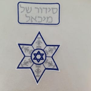 May include: Two embroidered patches. One is a blue and white Star of David with floral accents. The other is a rectangular patch with Hebrew text in gray. Both patches have a white background.