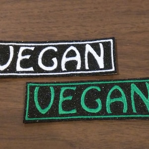Puede incluir: Dos parches rectangulares con la palabra "VEGAN" en letras blancas y verdes. Los parches tienen un fondo negro con purpurina. Un parche tiene un borde blanco y el otro un borde verde.