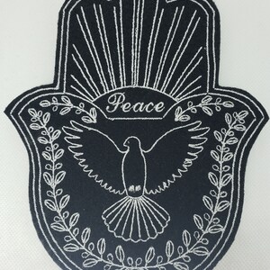 May include: A black felt Hamsa hand with white embroidery. The embroidery includes a dove with outstretched wings, a wreath of leaves, and the word "Peace".