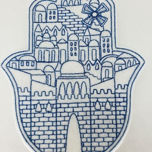 May include: Embroidered Hamsa hand patch with a detailed blue and white city scene, including buildings, domes, and a windmill. The patch has a denim-like texture and is likely for decorative purposes.