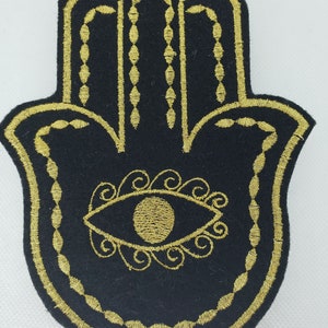 May include: A black felt patch with a gold embroidered Hamsa hand design. The Hamsa hand has an eye in the center and is surrounded by a decorative border.