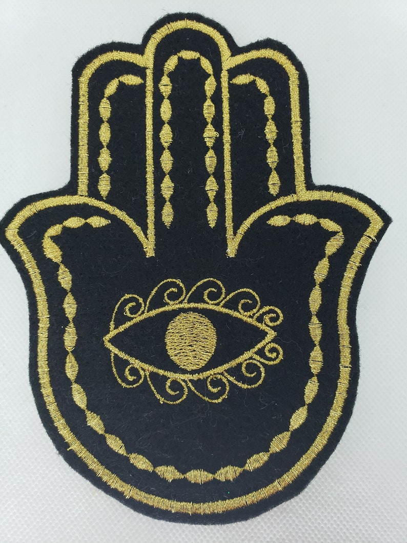 May include: A black felt patch with a gold embroidered Hamsa hand design. The Hamsa hand has an eye in the center and is surrounded by a decorative border.