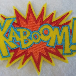 Puede incluir: Un parche de fieltro amarillo con una explosión roja y naranja detrás de la palabra "KA-BOOM!" en letras azules delineadas.