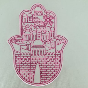 May include: A pink embroidered Hamsa hand patch featuring a detailed cityscape design. The patch includes buildings, arches, and a windmill, all outlined in pink thread. The background is a light, neutral color.