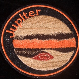 以下が含まれることがあります： オレンジと黒の刺繍が施された丸いパッチで、オレンジ色の文字で「Jupiter」と書いてあり、茶色とオレンジのストライプ模様が描かれています。