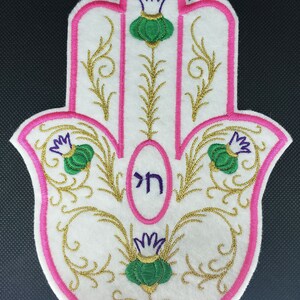 Könnte beinhalten: Eine weiße Hamsa-Hand mit rosa Umrandung und goldener Stickerei. Die Hamsa hat drei grüne und weiße Blumen mit violetten Zentren. Das Wort "Hamsa" ist in Hebräisch in der Mitte der Hamsa geschrieben.