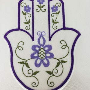 Puede incluir: Mano de Hamsa bordada en morado y verde con diseños florales. La mano de Hamsa es un símbolo de protección y buena suerte.