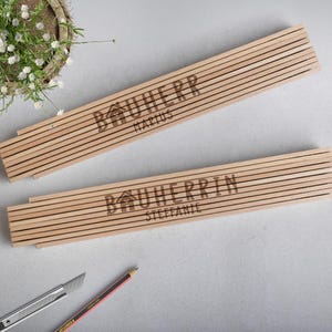 Könnte beinhalten: Zwei Holzlineale mit dem Text "Bauherrin" und den Namen "Marius" und "Stefanie" eingraviert.