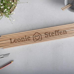 Könnte beinhalten: Ein Holzschild mit den Namen Leonie und Steffen und einem Haus mit einem Herzen darin. Das Schild besteht aus mehreren dünnen Holzlatten.