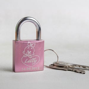Könnte beinhalten: Rosafarbener Vorhängeschloss mit einem silbernen Metallscharnier und einer personalisierten Inschrift mit dem Text "Lilly 14.09.2013" und einem Cartoon-Bild eines kleinen Mädchens in einem Herz.