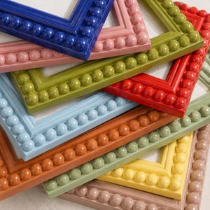 May include: A collection of stacked rectangular picture frames featuring a beaded border design. The frames are finished in a variety of solid colors including blue, pink, olive green, red, light blue, orange, sage green, yellow, and beige.
