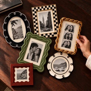 May include: A collection of six picture frames in various shapes and colors, including a black scalloped oval, a black and white checkered rectangle, a gold twisted rope rectangle, a green wavy rectangle, a red square, and a white scalloped circle.