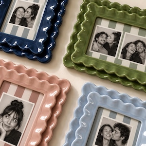 May include: Four rectangular ceramic picture frames with scalloped edges in glossy navy blue, olive green, soft pink, and light blue. Each frame contains a black and white photograph of people and features a striped background matting.