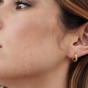 Peut inclure: Gros plan d'une boucle d'oreille dorée avec un motif de feuilles. La boucle d'oreille est portée à l'oreille d'une personne à la peau claire et aux cheveux bruns. Le bijou est petit et délicat.