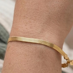 Peut inclure: Un bracelet en or à chevrons. Le bracelet est composé de petits maillons plats et imbriqués, créant une bande lisse et souple. Le bracelet est sécurisé par un petit fermoir et comporte un charm en forme de cœur. Le bracelet est porté sur un poignet.
