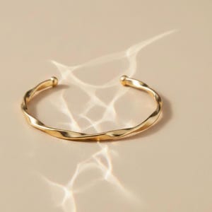 Peut inclure: Un bracelet manchette ouvert de couleur or avec un design torsadé. Le bracelet a une finition lisse et polie et des extrémités arrondies. Le bijou est présenté sur une surface de ton neutre avec des reflets de lumière.