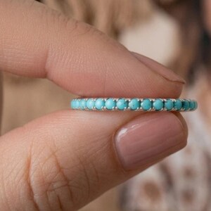 Peut inclure: Une bague en argent ornée d'une rangée de petites pierres rondes de couleur turquoise. La bague est tenue entre deux doigts, mettant en valeur la couleur bleu-vert vif des pierres. C'est un bijou.