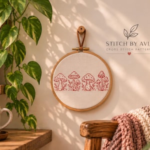 May include: A circular wooden embroidery hoop containing white fabric with a red cross-stitch pattern of four distinct mushroom clusters. The text STITCH BY AVIA CROSS STITCH PATTERNS is printed above the hoop in black with a small red heart.