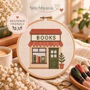 May include: A cross-stitch project in a wooden embroidery hoop featuring a bookshop storefront with a red awning, a green door, and the word BOOKS stitched in black. The design includes a window display with books and a potted plant beside the door.