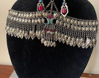 Puede incluir: Juego de cabeza y pendientes plateados con detalles rojos y turquesas. La pieza para la cabeza presenta una banda ancha con colgantes en forma de hoja y un colgante central. Los pendientes tienen forma de lágrima con una piedra roja y detalles turquesas.