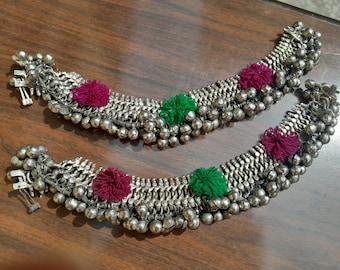May include: Two silver anklets with small bells and decorative accents. Each anklet features a series of silver bells and is adorned with three colorful pom-poms in shades of green and burgundy. The anklets are displayed on a wooden surface.
