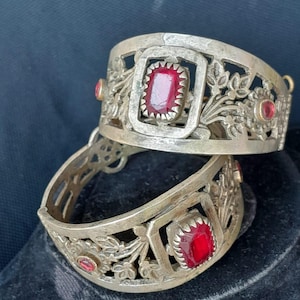 Può includere: Due braccialetti color argento con intricati ritagli floreali e geometrici. Ogni braccialetto presenta una grande gemma rossa rettangolare al centro, circondata da piccoli accenti rossi. I braccialetti sono esposti su una superficie scura.