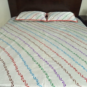 May include: White bed linen set featuring a colourful, embroidered, wavy pattern. The design includes lines of red, blue, green, purple, and brown. The pillowcases have a red border.