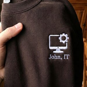 May include: A brown sweatshirt with a white embroidered design of a computer monitor and gear. The text "John, IT" is embroidered below the graphic. The sweatshirt is folded and held by a hand.