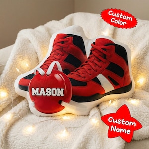 May include: Red and black high-top sneakers with white accents are displayed on a soft, white blanket. A red ornament with the name "MASON" is in front of the sneakers. A red star-shaped tag says "Custom Name". A red oval-shaped tag says "Custom Color".