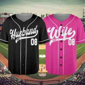 May include: Two baseball jerseys, one black and one pink, displayed against a baseball field backdrop. The black jersey says "Husband" with "2025" and "08" in white. The pink jersey says "Wife" with "2025" and "08" in white.