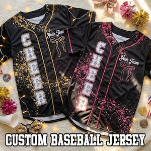 May include: Two black custom baseball jerseys with the word "CHEER" in white block letters. One jersey has gold accents and the other has pink accents. Both jerseys have the text "Your Team" and a bow graphic. The text "CUSTOM BASEBALL JERSEY" is at the bottom.