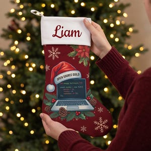 May include: A deep red Christmas stocking with a white cuff personalized with the name Liam. The stocking features a graphic of a laptop wearing a Santa hat, surrounded by pine branches, pinecones, poinsettias, and snowflakes.