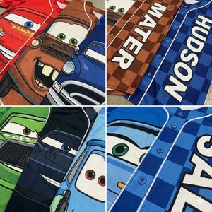 May include: Baseball jerseys with cartoon car designs in red, brown, blue, and green. The jerseys have button-up fronts and include character names like "Mater" and "Hudson" in white block letters.