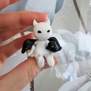 May include: A small, handmade figurine of a white devil-like character with black wings and a black creature in its arms. The figurine has small horns, black eyes, and a minimalist design. The figure is held in a person's hand.